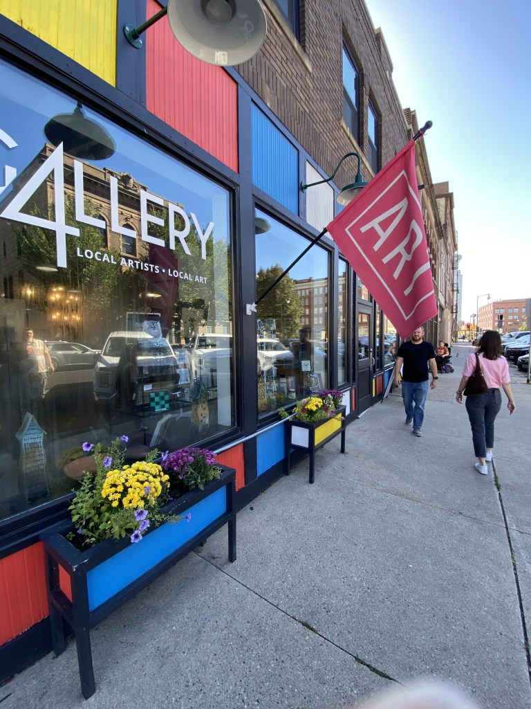 Exterior view of a colorful art gallery showcasing local artists, with a prominent 'ART' flag, flowers in planters, and pedestrians walking by.