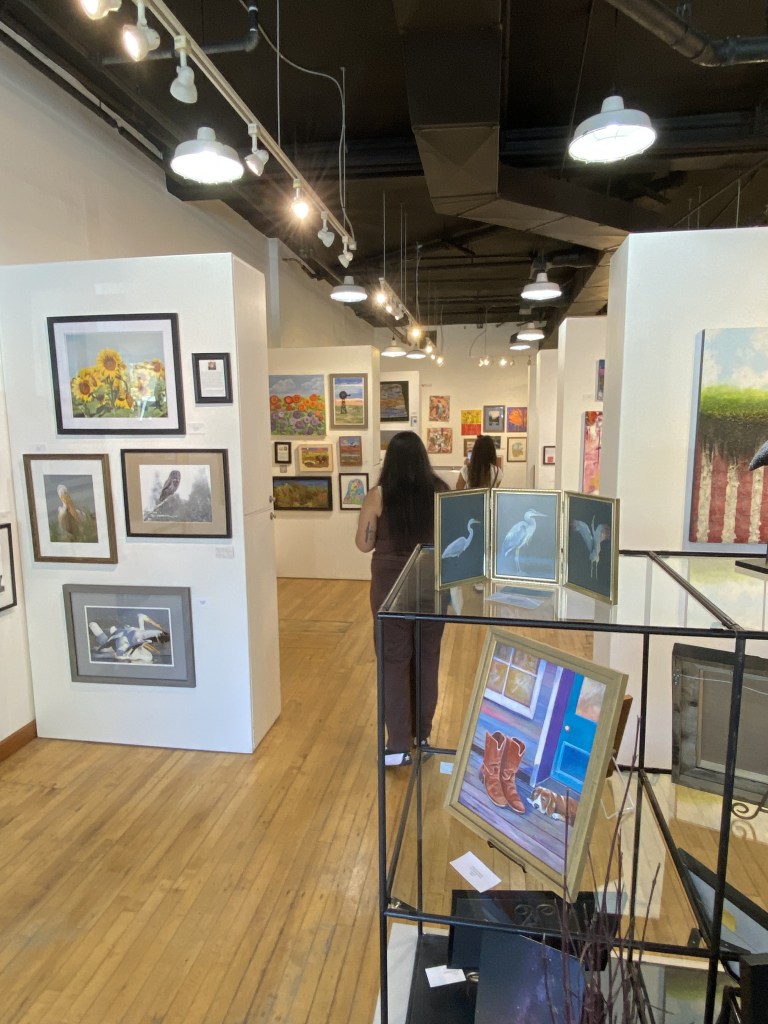 Art gallery interior featuring various paintings on white walls, with two visitors admiring the artwork. A display case in the foreground holds framed art and decorative items.