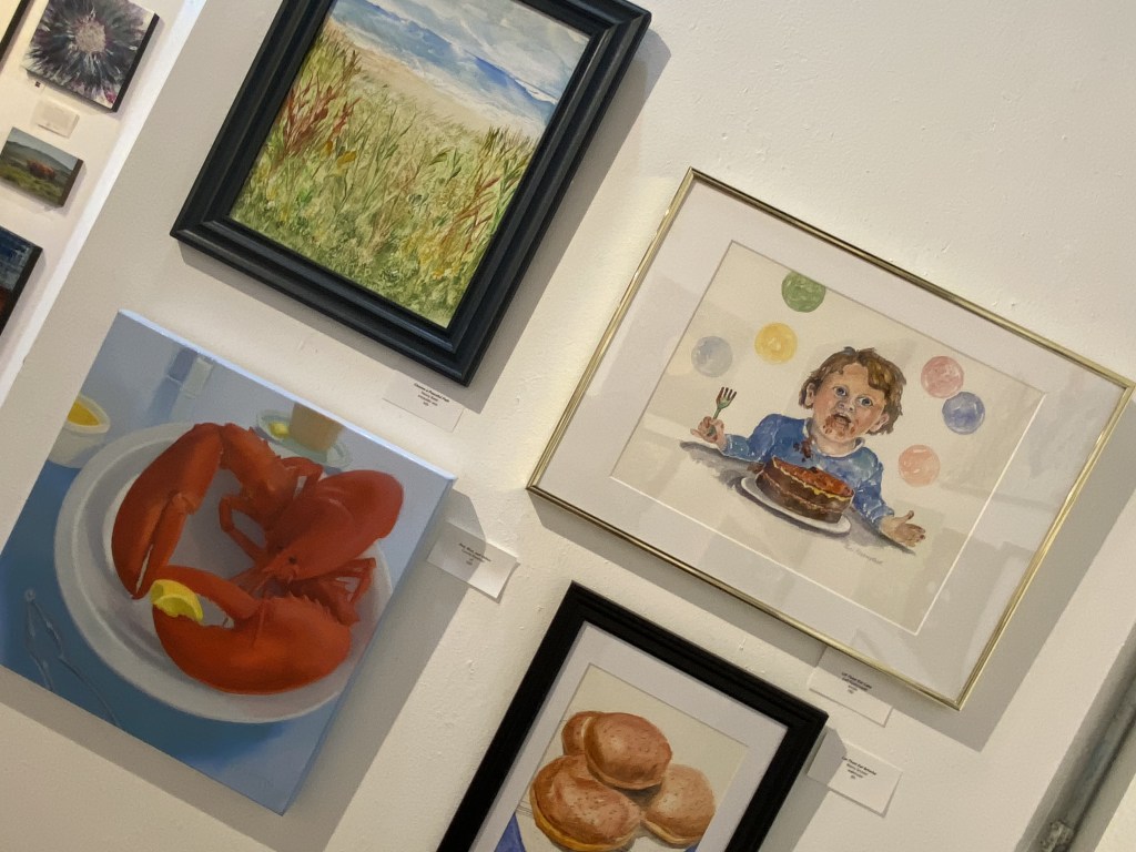 A collection of paintings on a gallery wall, featuring a landscape of grass and ocean, a vibrant depiction of lobsters on a plate, a child with a fork looking surprised at a dessert, and a still life of freshly baked rolls.