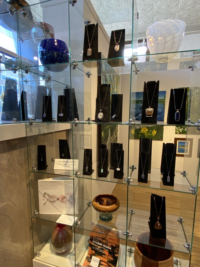 Display case showcasing various necklaces on black stands with a variety of designs, including pendants. The background contains decorative glass sculptures and artwork.