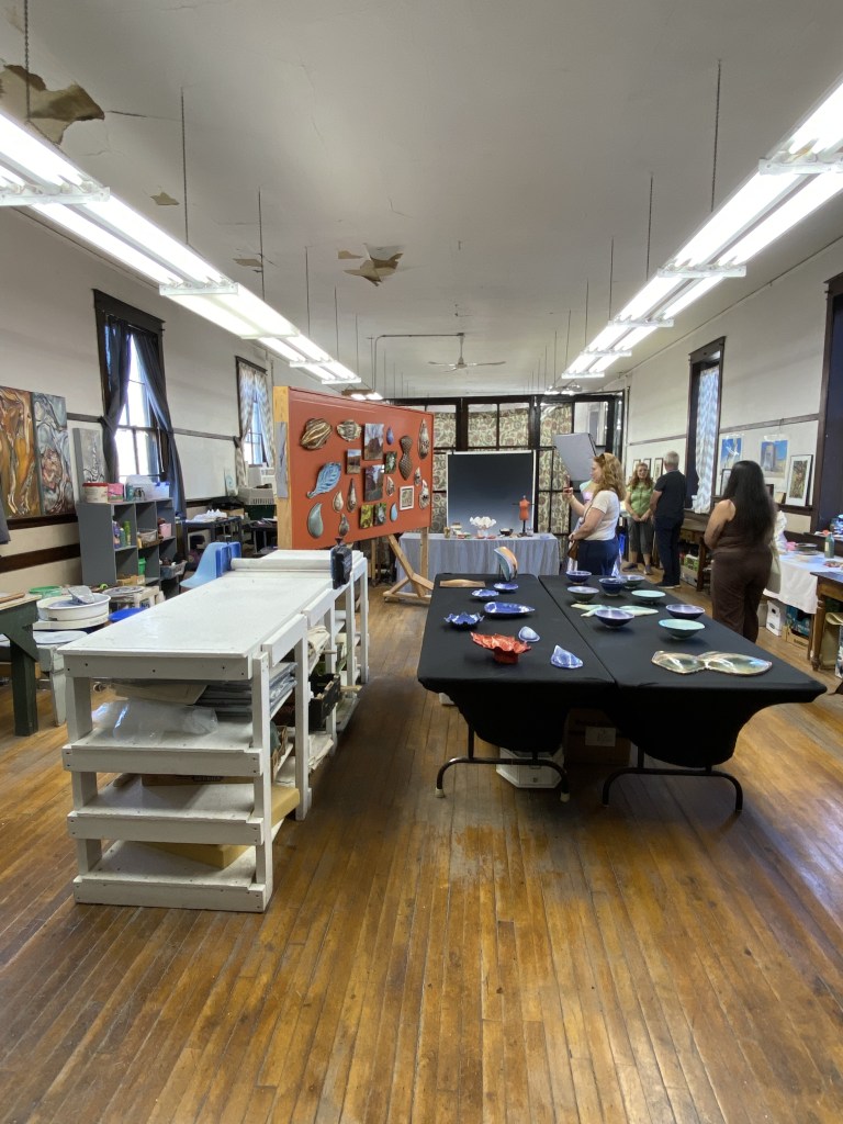Interior view of an art studio featuring wooden floors, tall windows, and fluorescent lighting. A black table is set up with colorful plates, and an orange wall displays various artworks. People are seen engaging in activities around the studio.