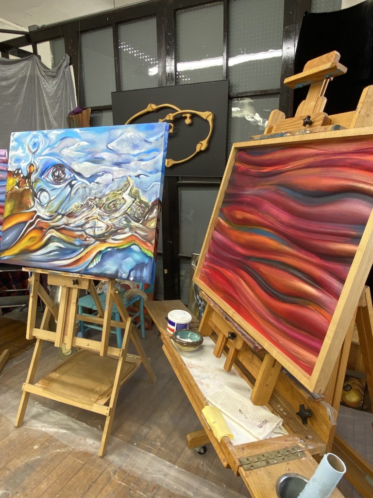 An artist's studio featuring two canvases on easels: one with abstract blue and yellow patterns resembling waves, and the other with rich red and orange flowing lines.