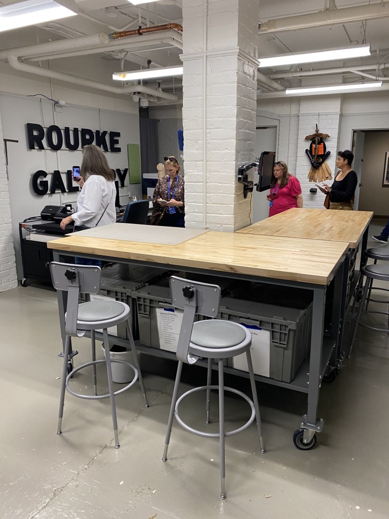 Interior view of the Rourke Gallery showcasing a workspace with stools, a large wooden table, and people engaging in conversation.