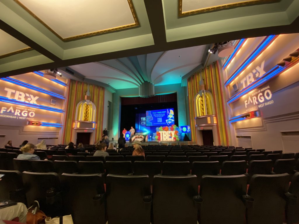 Interior view of a theater set up for an event, featuring a stage with colorful decor and seating area with empty chairs.