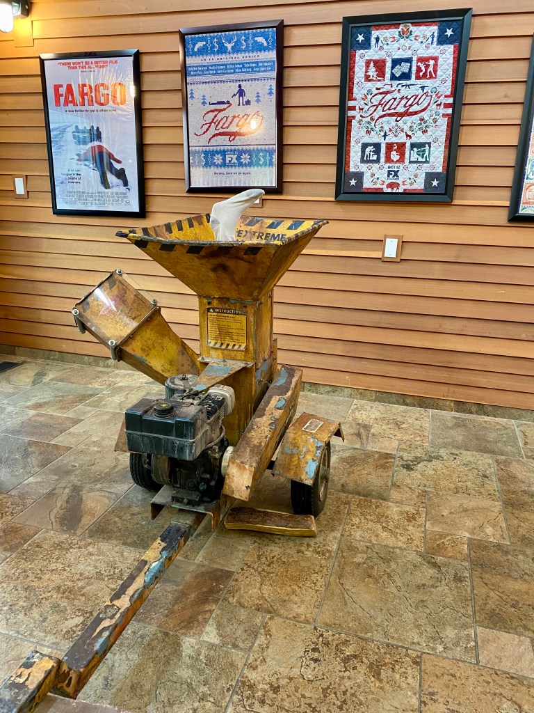 A vintage wood chipper on display in a gallery, featuring a yellow and black exterior with a powerful engine, set against a backdrop of Fargo-themed artwork.